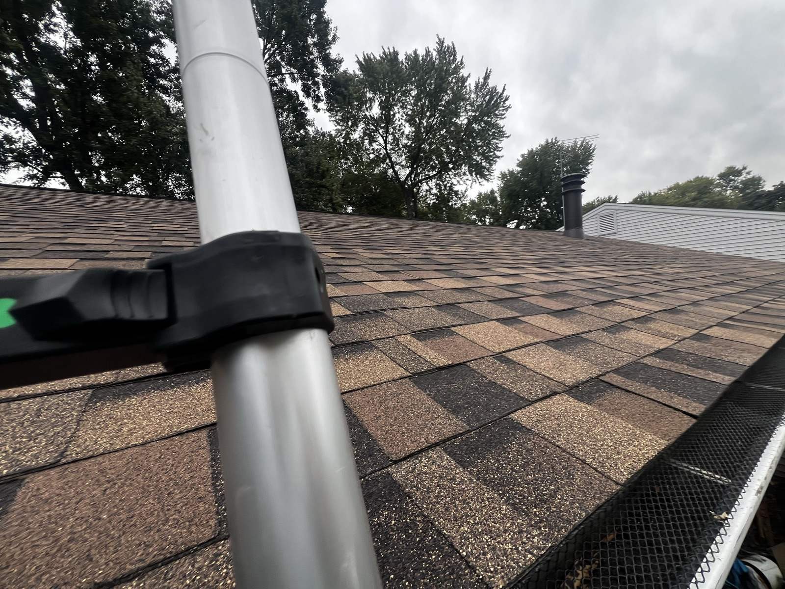 A ladder rail with a black standoff bracket clamped to it, leaning across a run of new brown and tan shingles near the eave, with perforated gutter guard visible along the trough at the lower right and trees under an overcast sky