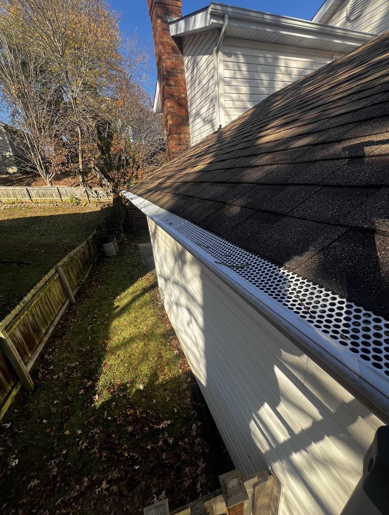 Perforated aluminium gutter guard running along an eave of dark shingles, with a red brick chimney and white lap siding above and a fenced back garden covered in fallen leaves below
