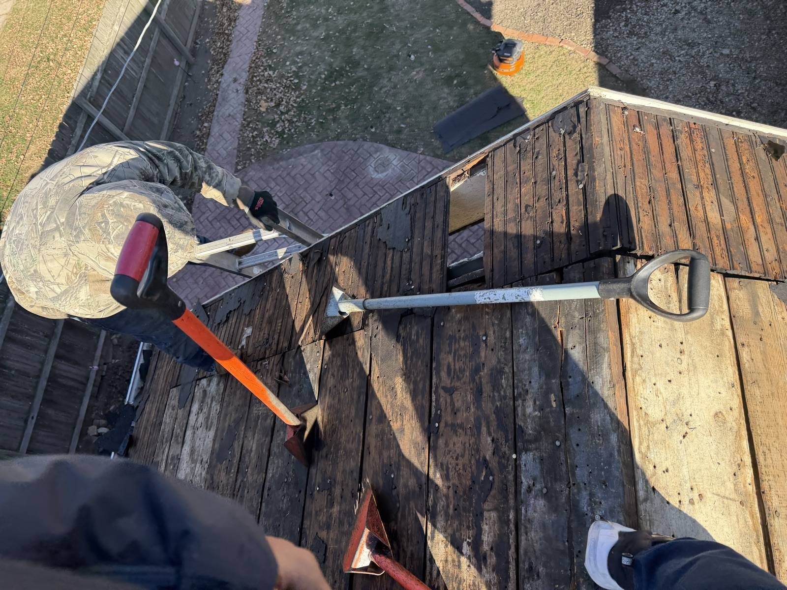 Looking straight down at two long handled tear off spades and a flat pry bar lying on a roof stripped to bare board decking, with a worker in a camouflage hoodie kneeling at the far edge and a brick paved patio on the ground below
